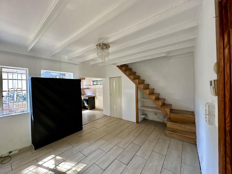 Open plan living area featuring tiled floors, exposed beam ceilings, and a wooden staircase leading to an upper level, with an adjacent kitchen area.