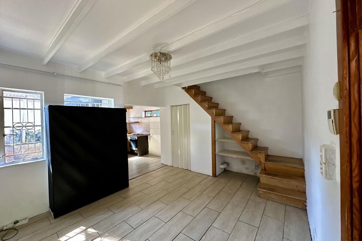 Open plan living area featuring tiled floors, exposed beam ceilings, and a wooden staircase leading to an upper level, with an adjacent kitchen area.