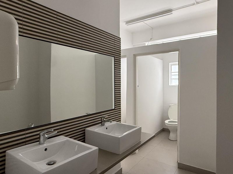 Modern office bathroom featuring a double vanity with vessel sinks, large wall mirror, and a separate toilet cubicle with tiled finishes.