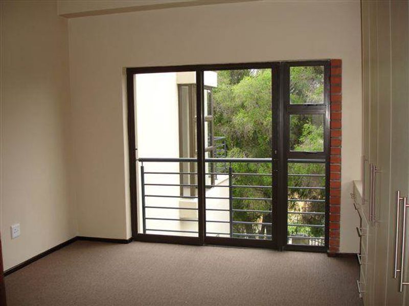 A vacant bedroom featuring built-in cupboards, carpeted flooring, and a sliding glass door opening onto a balcony with a view of surrounding trees.