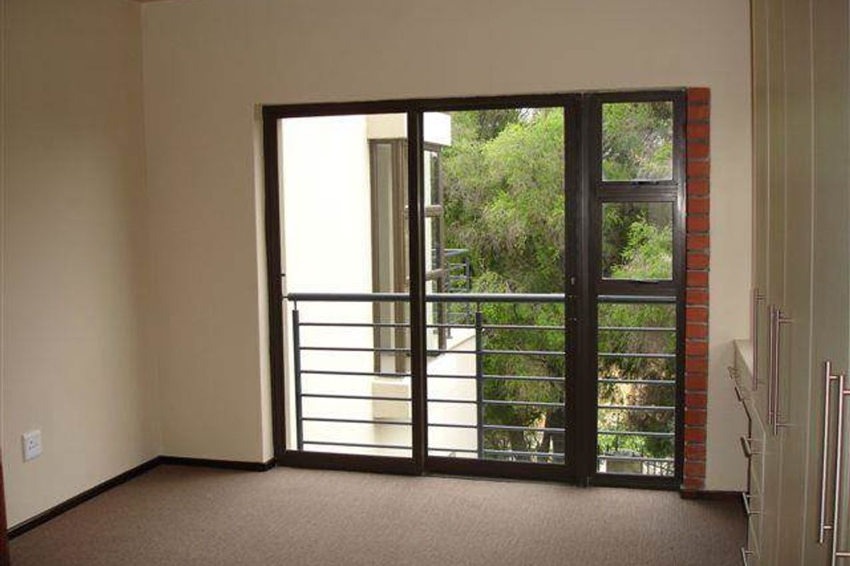 A vacant bedroom featuring built-in cupboards, carpeted flooring, and a sliding glass door opening onto a balcony with a view of surrounding trees.