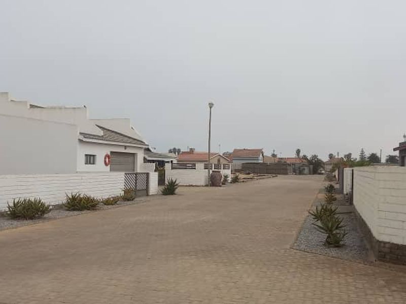A street view showing the paved road, surrounding residential properties, and boundary walls in a quiet neighborhood setting.