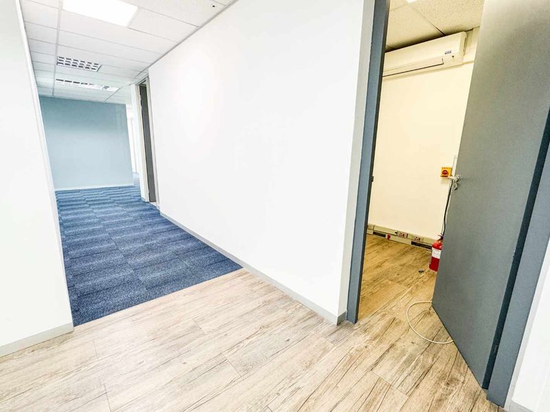 A clean, vacant office hallway featuring a mix of carpet and wood-look vinyl flooring, suspended ceilings, and an integrated air conditioning unit.