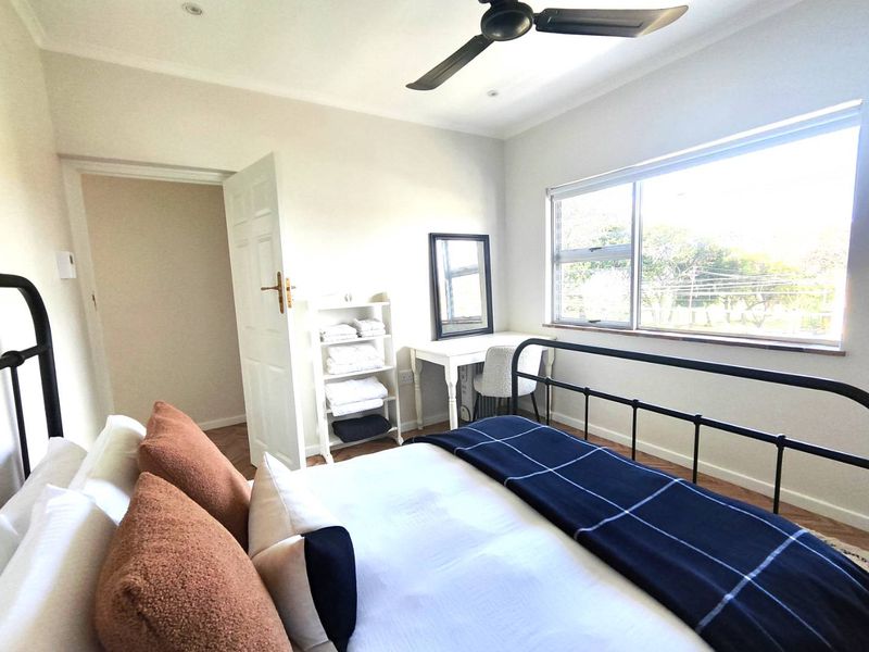 A bright bedroom featuring timber flooring, a ceiling fan, and large windows providing natural light.