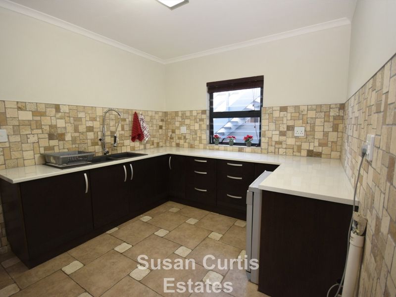 A functional kitchen featuring dark cabinetry, light stone-look countertops, and matching tiled splashbacks with a double sink and window view.