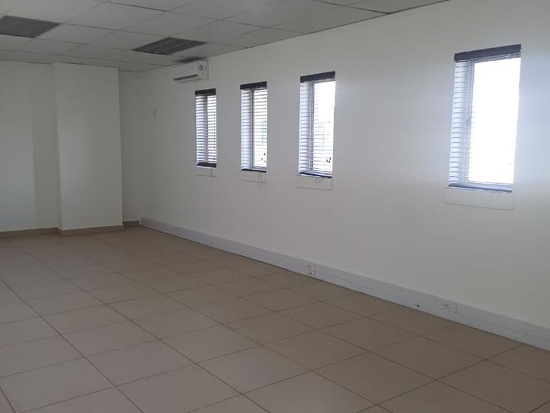 A spacious, empty office area featuring tiled flooring, a suspended ceiling with recessed lighting, and a wall-mounted air conditioning unit.