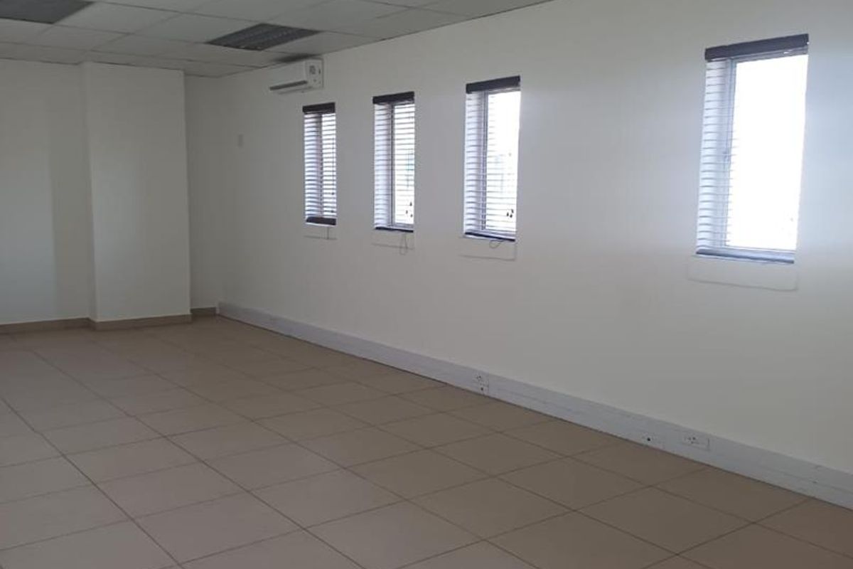 A spacious, empty office area featuring tiled flooring, a suspended ceiling with recessed lighting, and a wall-mounted air conditioning unit.