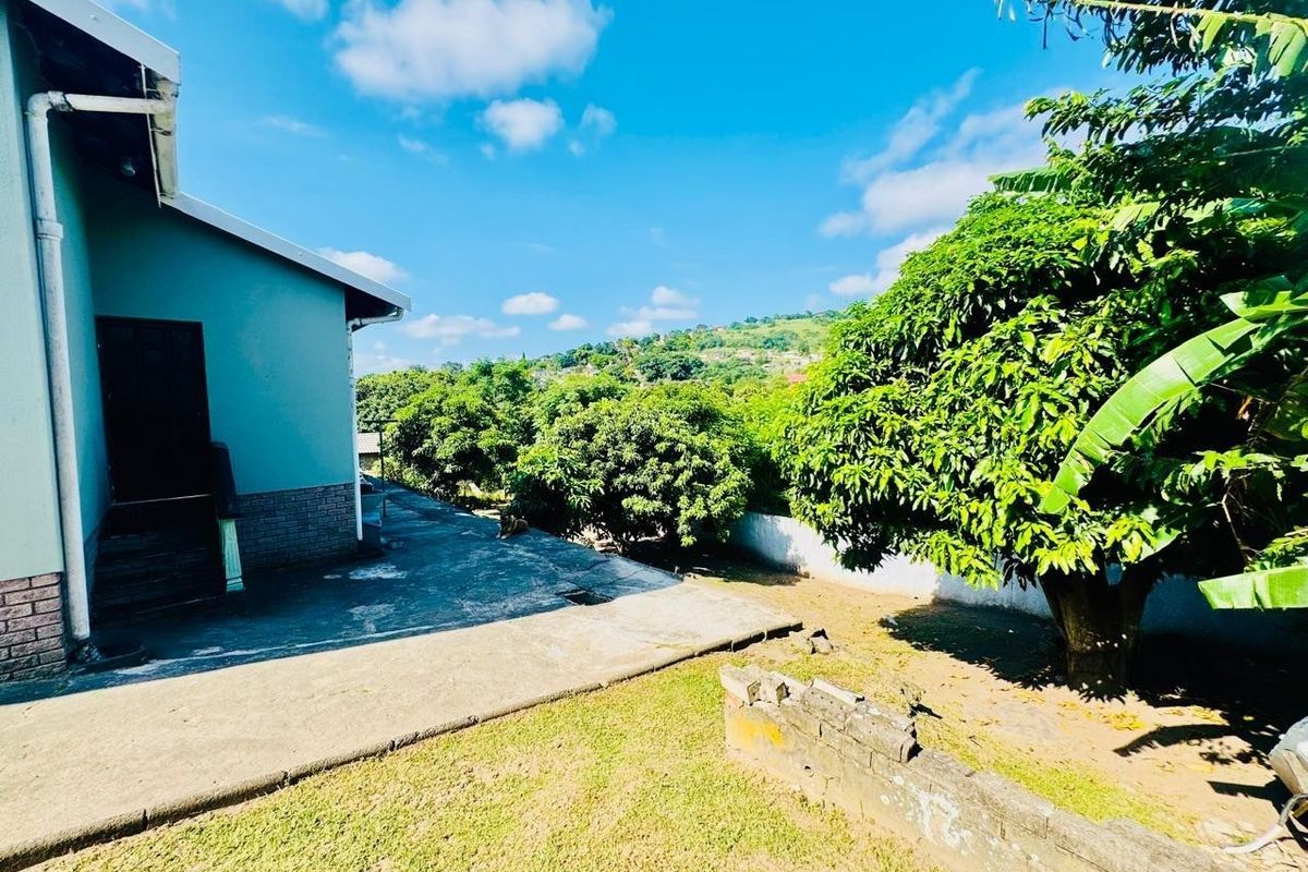 Rear exterior view of the property featuring a concrete patio area, mature trees, and a boundary wall with scenic hillside views.