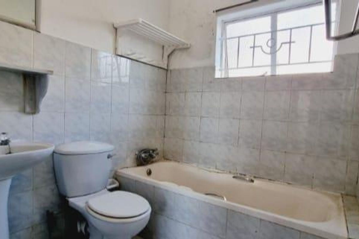 A dated bathroom featuring a standard bathtub, pedestal sink, and toilet, with wall-to-wall tiling and a security-grilled window.