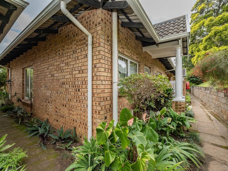 Side view of a face brick townhouse featuring a tiled roof, exposed rafters, and a paved walkway surrounded by established garden beds.