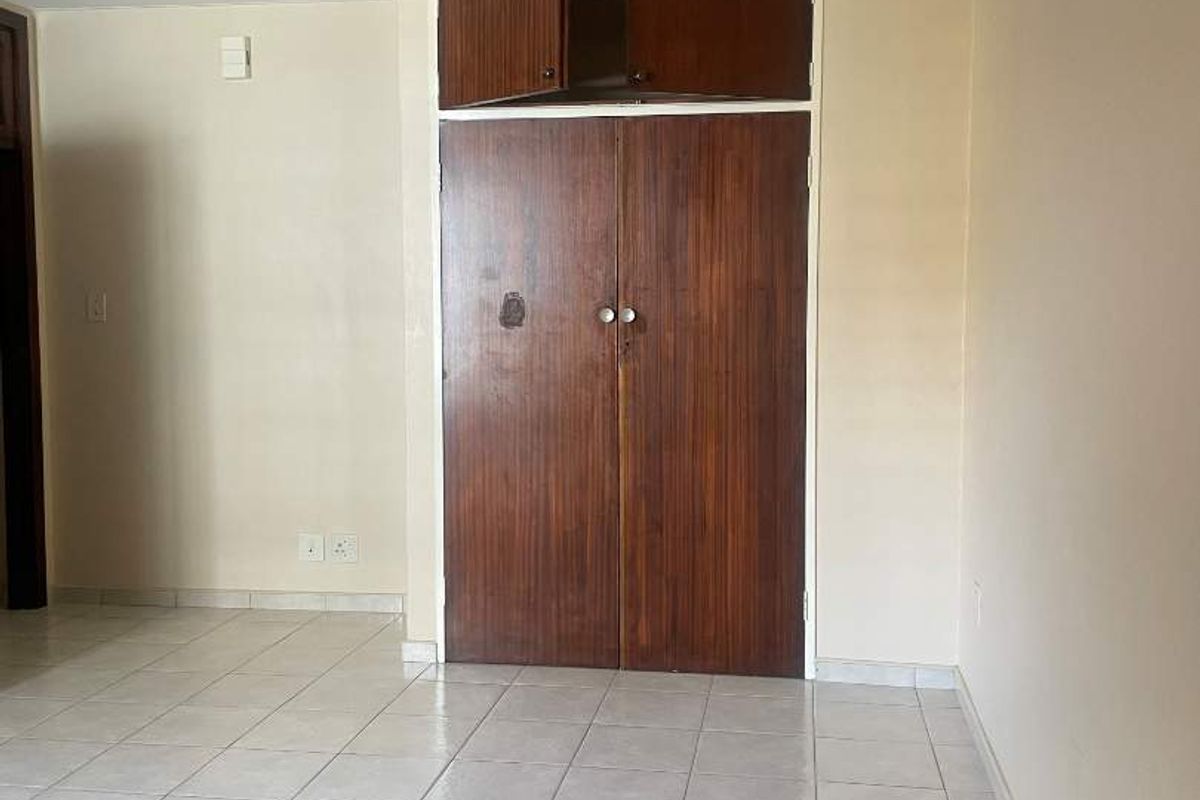 An empty bedroom featuring tiled flooring and a dated built-in wooden wardrobe with overhead storage compartments.