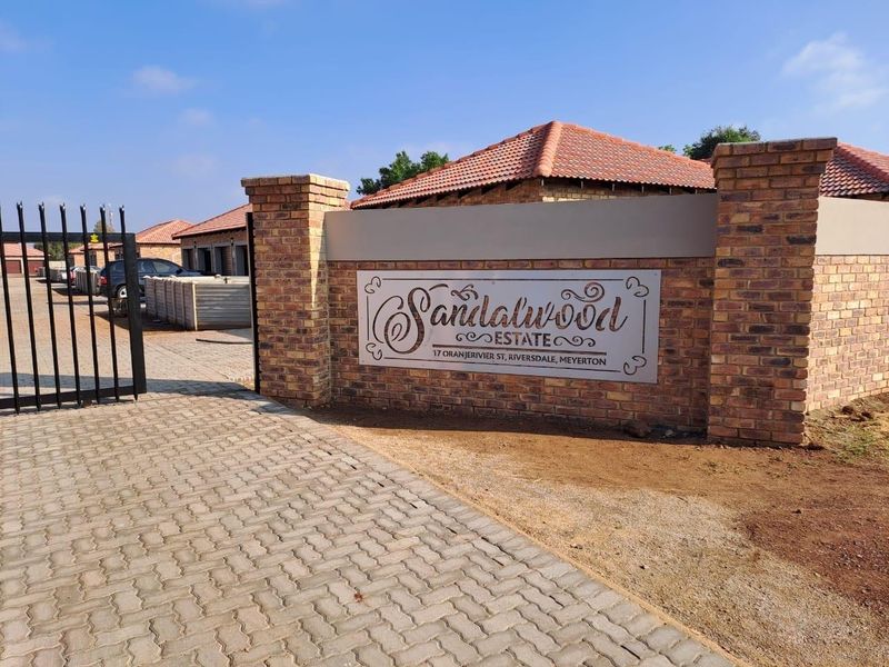 Entrance view of Sandalwood Estate featuring a brick boundary wall, paved driveway, and secure metal gate access.