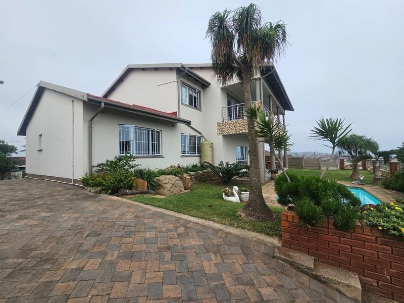 Rear view of a multi-story house featuring a paved driveway, swimming pool, balcony with stone detailing, and established garden area.