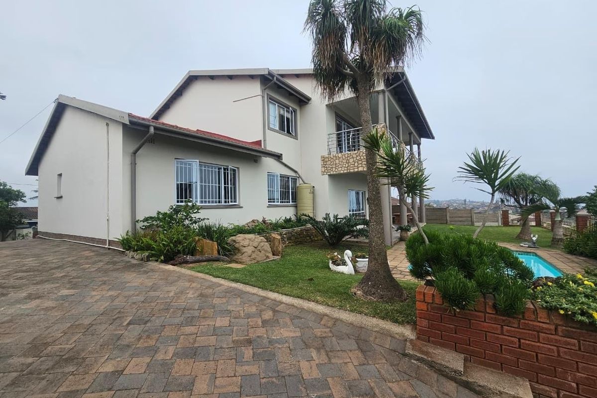 Rear view of a multi-story house featuring a paved driveway, swimming pool, balcony with stone detailing, and established garden area.