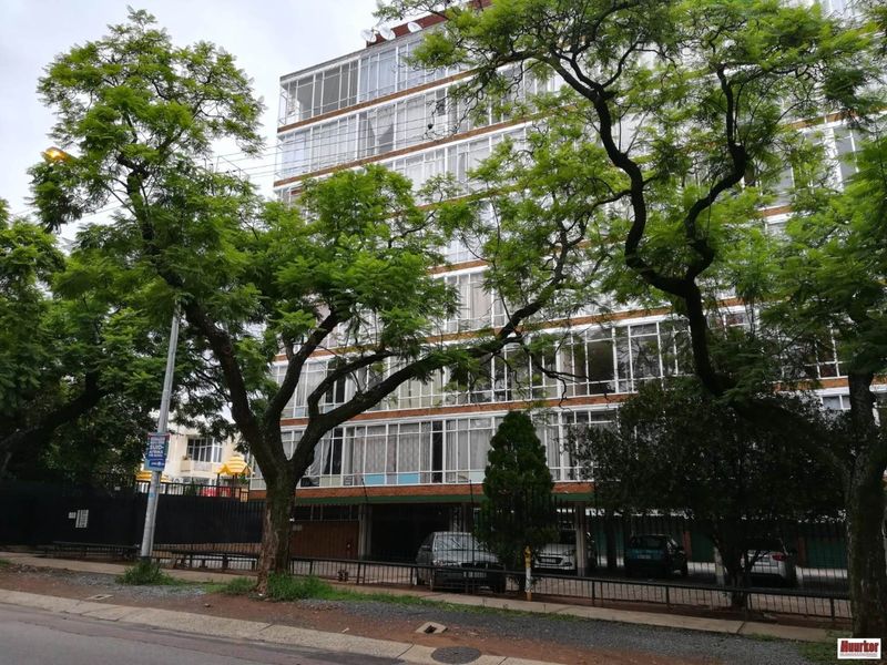 Street view of a multi-story apartment building featuring brick facade, enclosed balconies, and ground-level parking facilities.