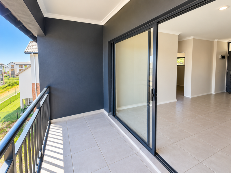 A modern, covered balcony featuring neutral tiled flooring, a sleek metal balustrade, and large sliding glass doors leading into an open-plan interior space.