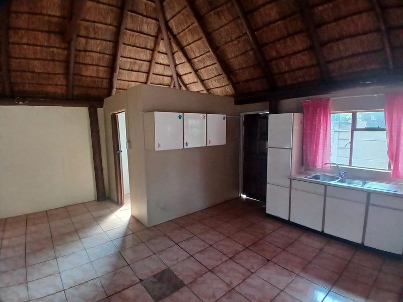 Open plan living area featuring a traditional thatched roof, exposed timber beams, tiled flooring, and a functional kitchen kitchenette.