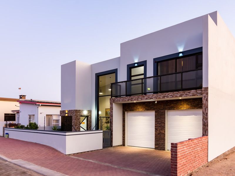 Modern double-story residence featuring a double garage, stone-clad accents, and an upper-level balcony with glass balustrades, set behind a secure boundary wall.