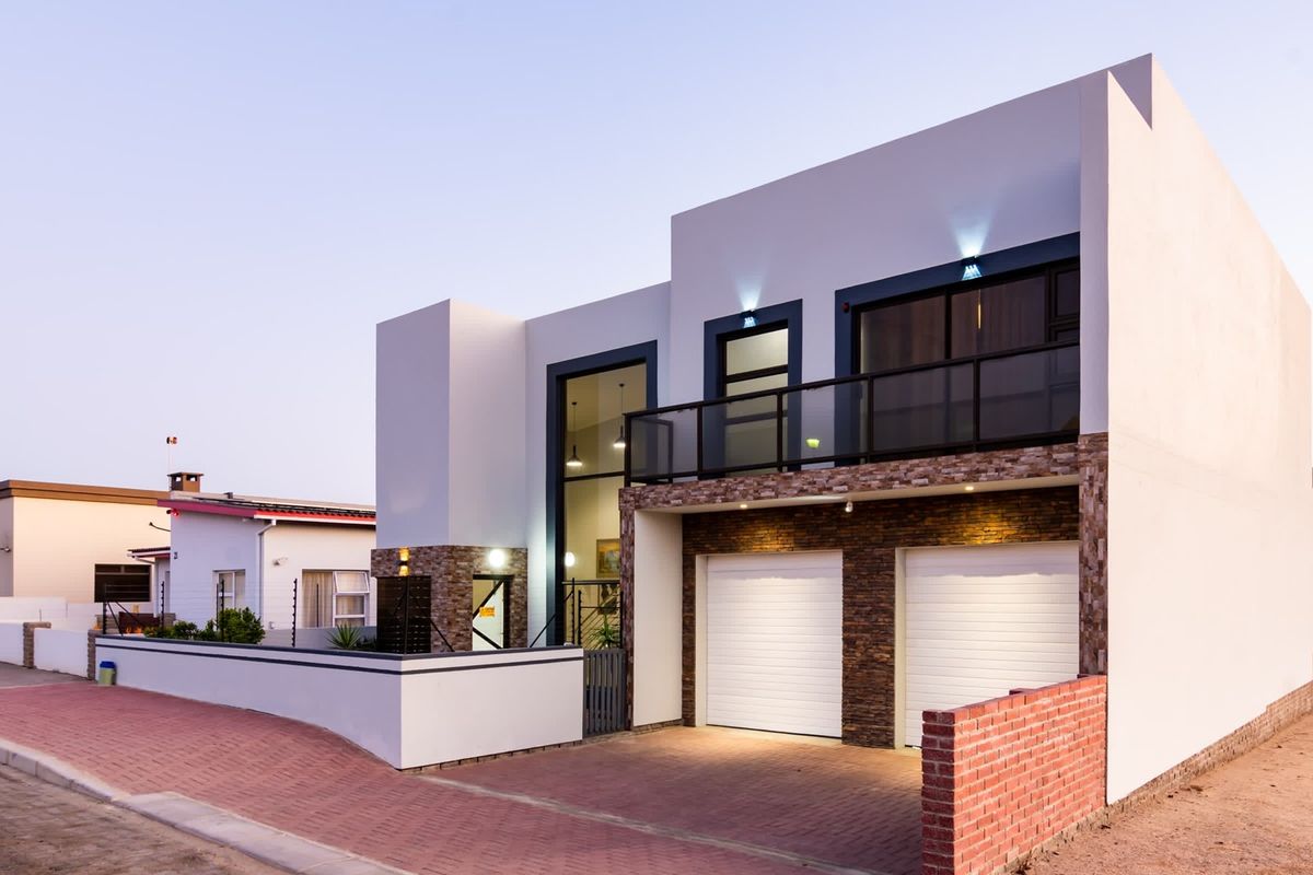 Modern double-story residence featuring a double garage, stone-clad accents, and an upper-level balcony with glass balustrades, set behind a secure boundary wall.