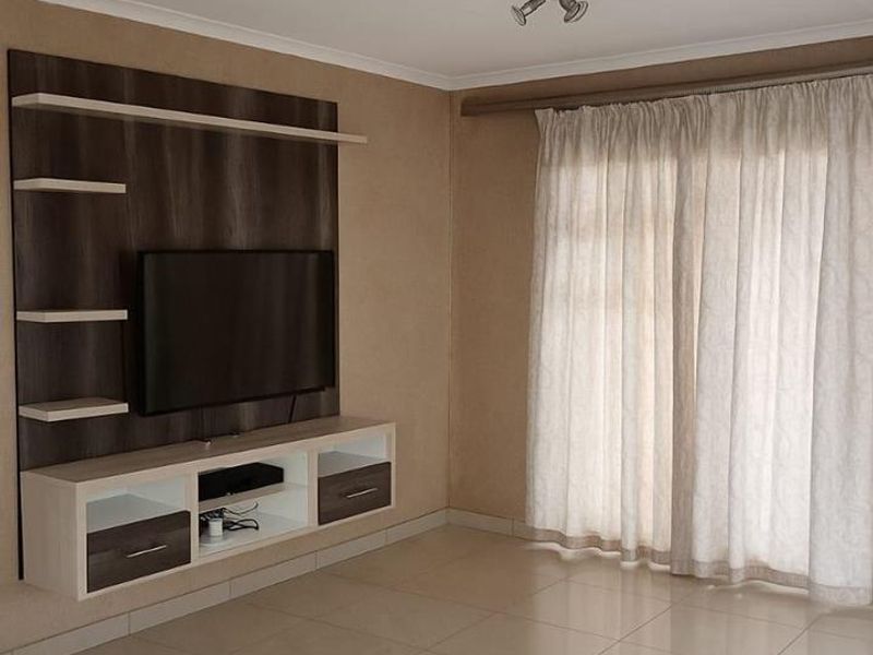 A bright and spacious living room featuring neutral tiled flooring, a modern built-in media unit, and large windows with installed curtain tracks.