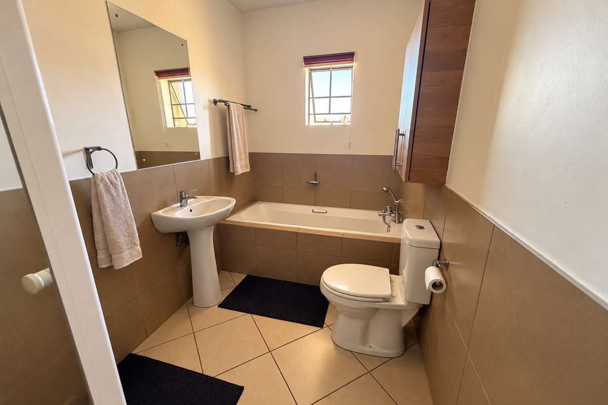 A functional bathroom featuring a pedestal basin, built-in bathtub, toilet, and wall-mounted storage cabinet with neutral tiled finishes.