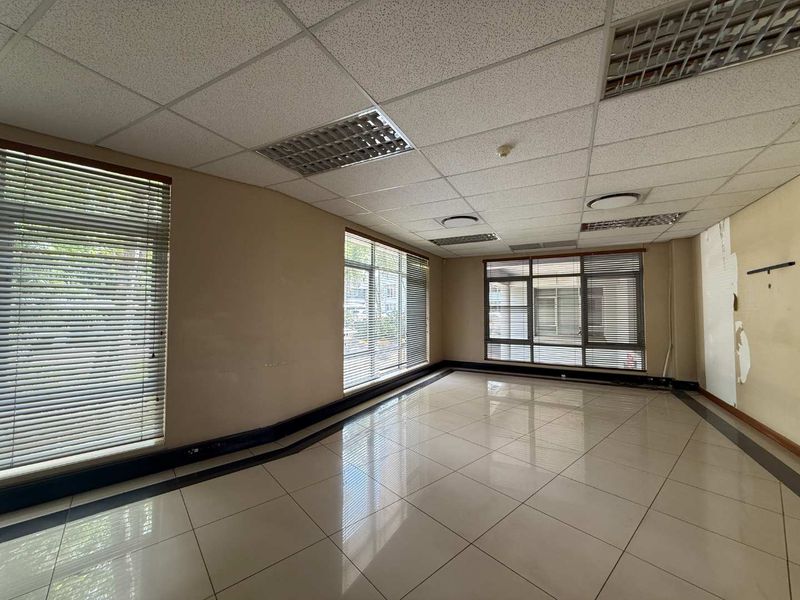 An empty office space featuring tiled flooring, a suspended ceiling with recessed lighting, and large windows with venetian blinds.