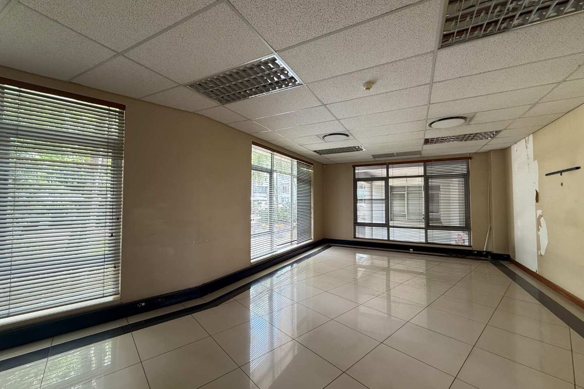 An empty office space featuring tiled flooring, a suspended ceiling with recessed lighting, and large windows with venetian blinds.