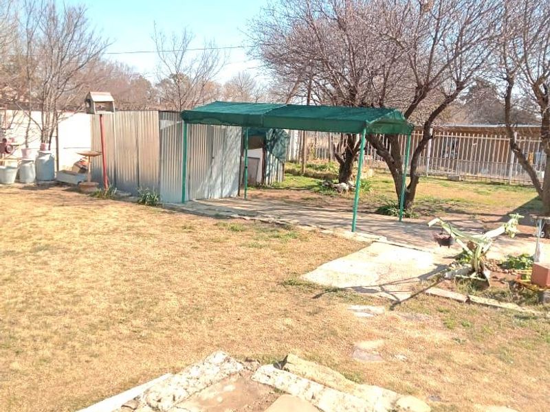 A spacious backyard featuring a corrugated metal shed, a shaded outdoor area, stone paving, and established trees within a fenced perimeter.