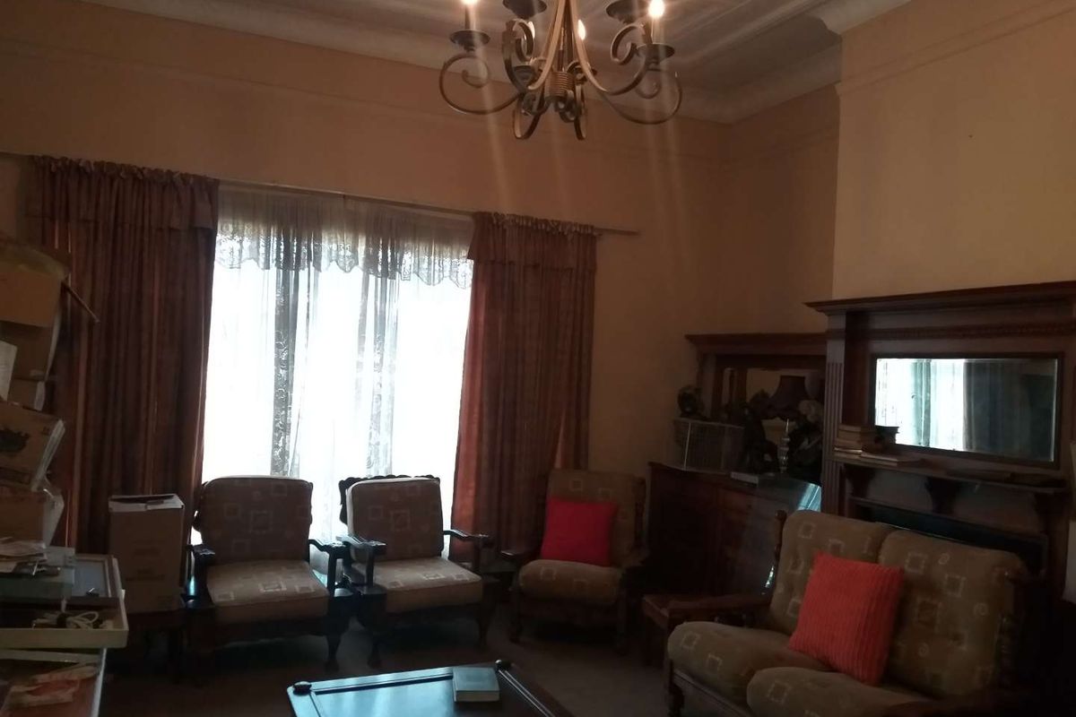 A traditional living room featuring a decorative ceiling rose, a built-in wooden fireplace mantel with mirror, and large windows with heavy drapery.