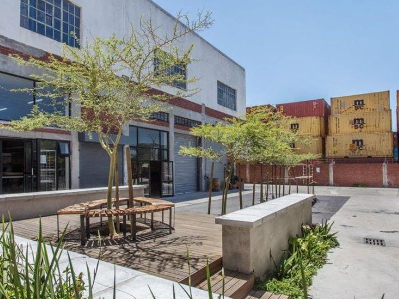 An industrial-style courtyard featuring timber decking, concrete seating, and direct access to the building via large steel-framed windows and roller shutter doors.