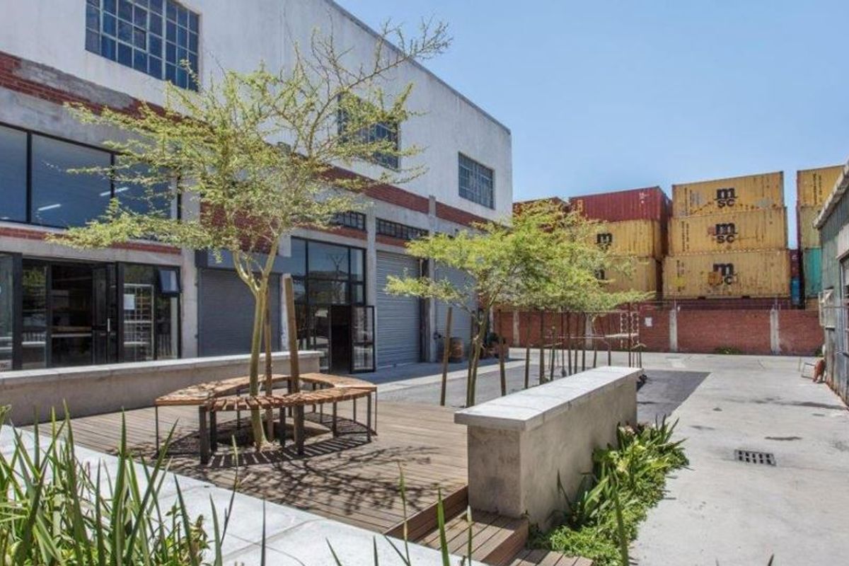 An industrial-style courtyard featuring timber decking, concrete seating, and direct access to the building via large steel-framed windows and roller shutter doors.