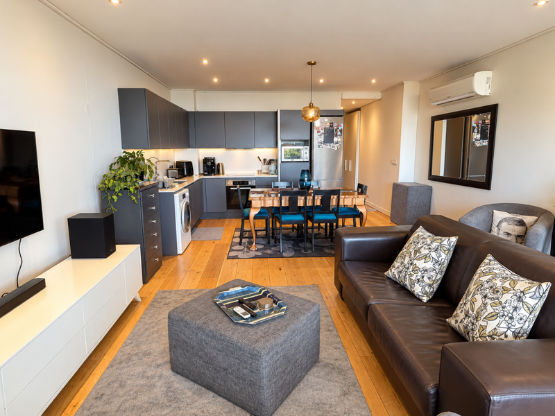 A modern open-plan living and kitchen area featuring dark grey cabinetry, integrated appliances, hardwood flooring, and a wall-mounted air conditioning unit.