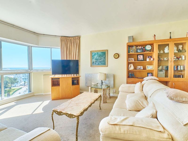 A bright living room featuring large floor-to-ceiling windows with expansive coastal views, carpeted flooring, and a built-in wooden shelving unit.