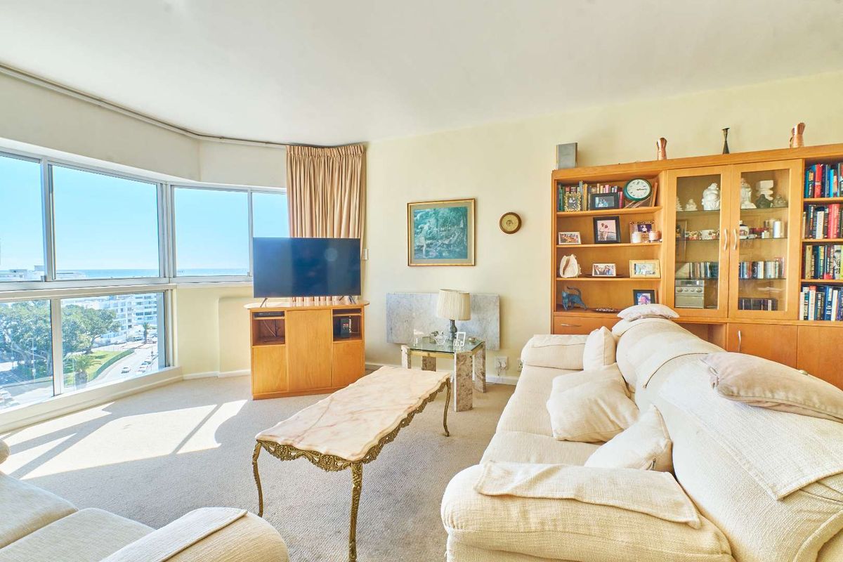 A bright living room featuring large floor-to-ceiling windows with expansive coastal views, carpeted flooring, and a built-in wooden shelving unit.
