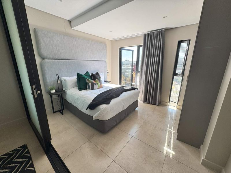 Modern bedroom featuring a large upholstered headboard, tiled flooring, recessed ceiling lighting, and a sliding glass door leading to a balcony.