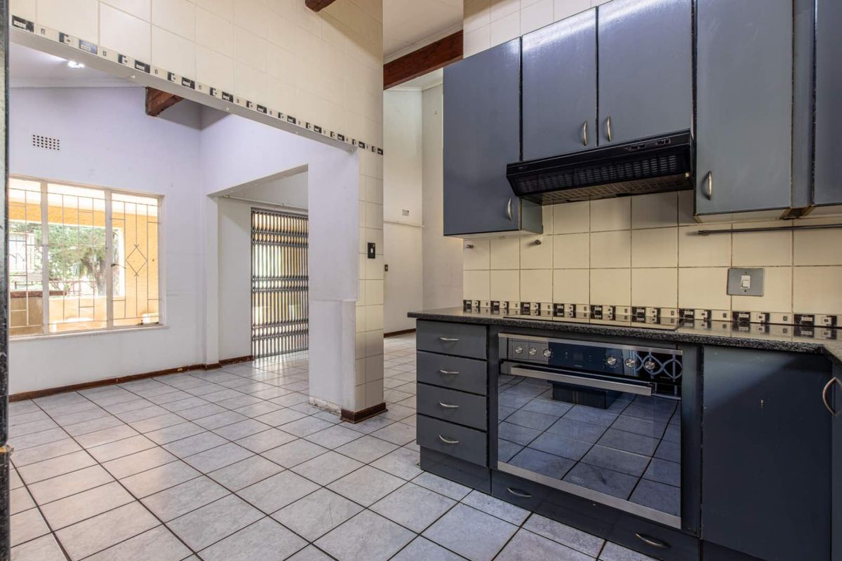 A functional kitchen featuring dark cabinetry, built-in oven, extractor fan, and tiled flooring, opening into an adjacent living area with security gates.