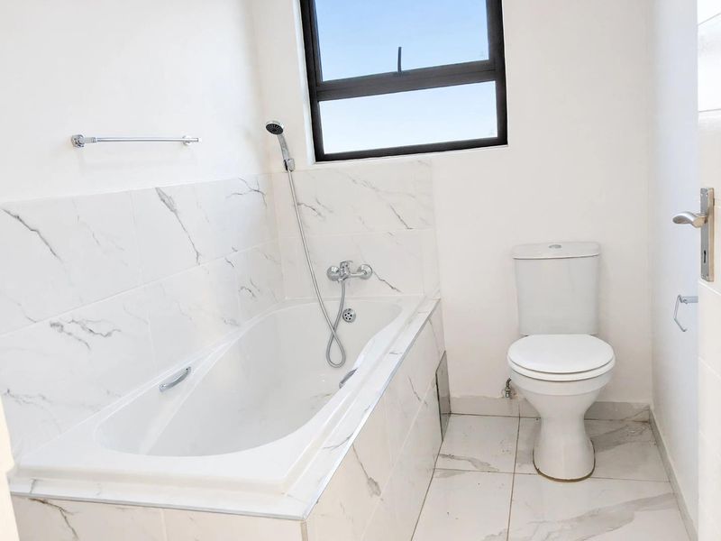 A clean, modern bathroom featuring white marble-look tiled walls and flooring, a built-in bathtub with a shower mixer, and a standard toilet.