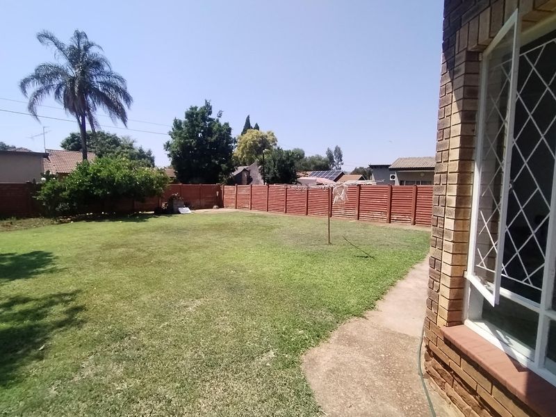 A spacious rear garden featuring a well-maintained lawn, brick boundary walls, and a concrete pathway leading along the side of the brick house.