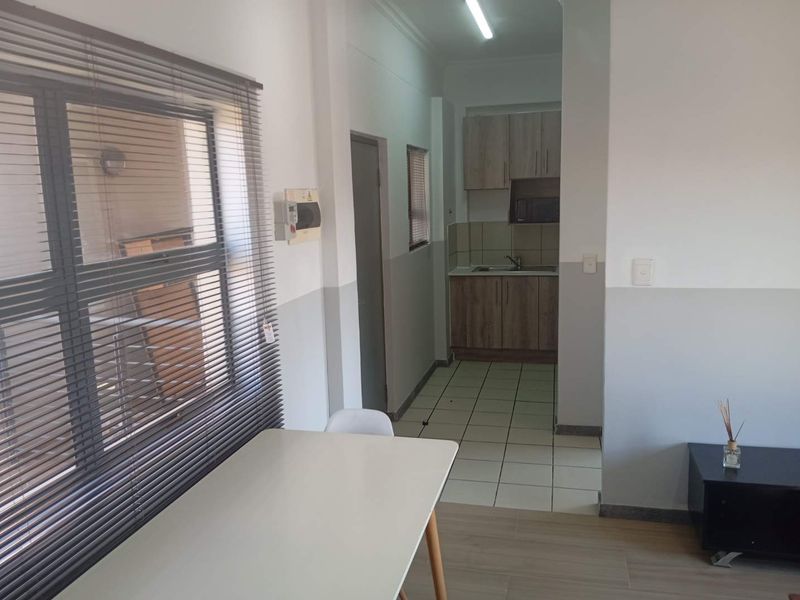 Open plan living area featuring a compact kitchen with wood-look cabinetry, tiled flooring, and large windows with venetian blinds.