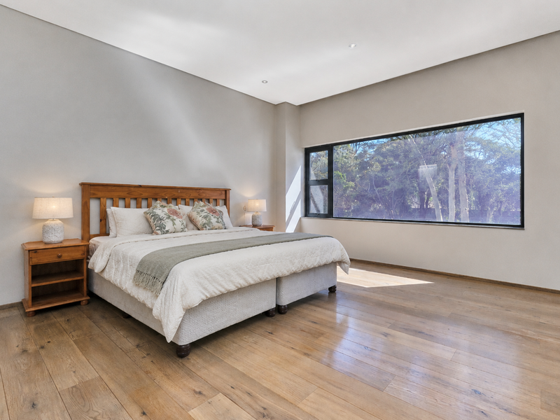 Spacious bedroom featuring high ceilings, recessed downlights, warm timber flooring, and a large window providing natural light and garden views.