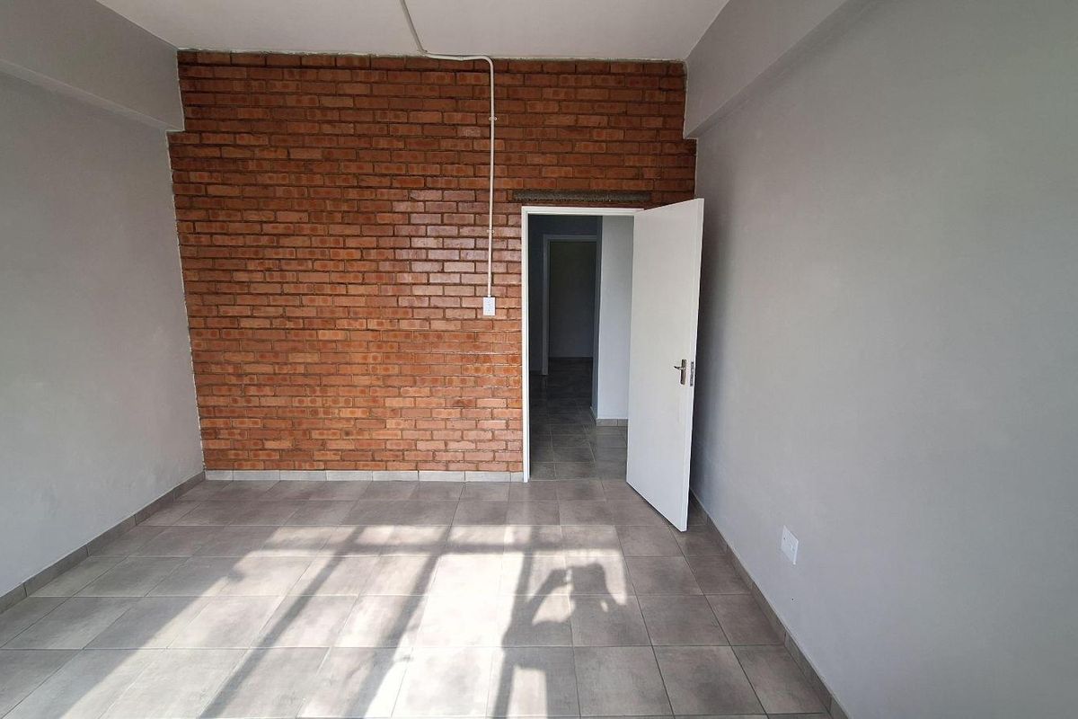 An empty bedroom featuring an exposed brick accent wall, neutral grey painted walls, and ceramic floor tiling.