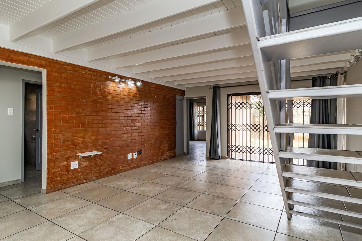 Spacious living area featuring an exposed brick feature wall, white exposed beam ceiling, tiled flooring, and a modern metal staircase leading to the upper level.