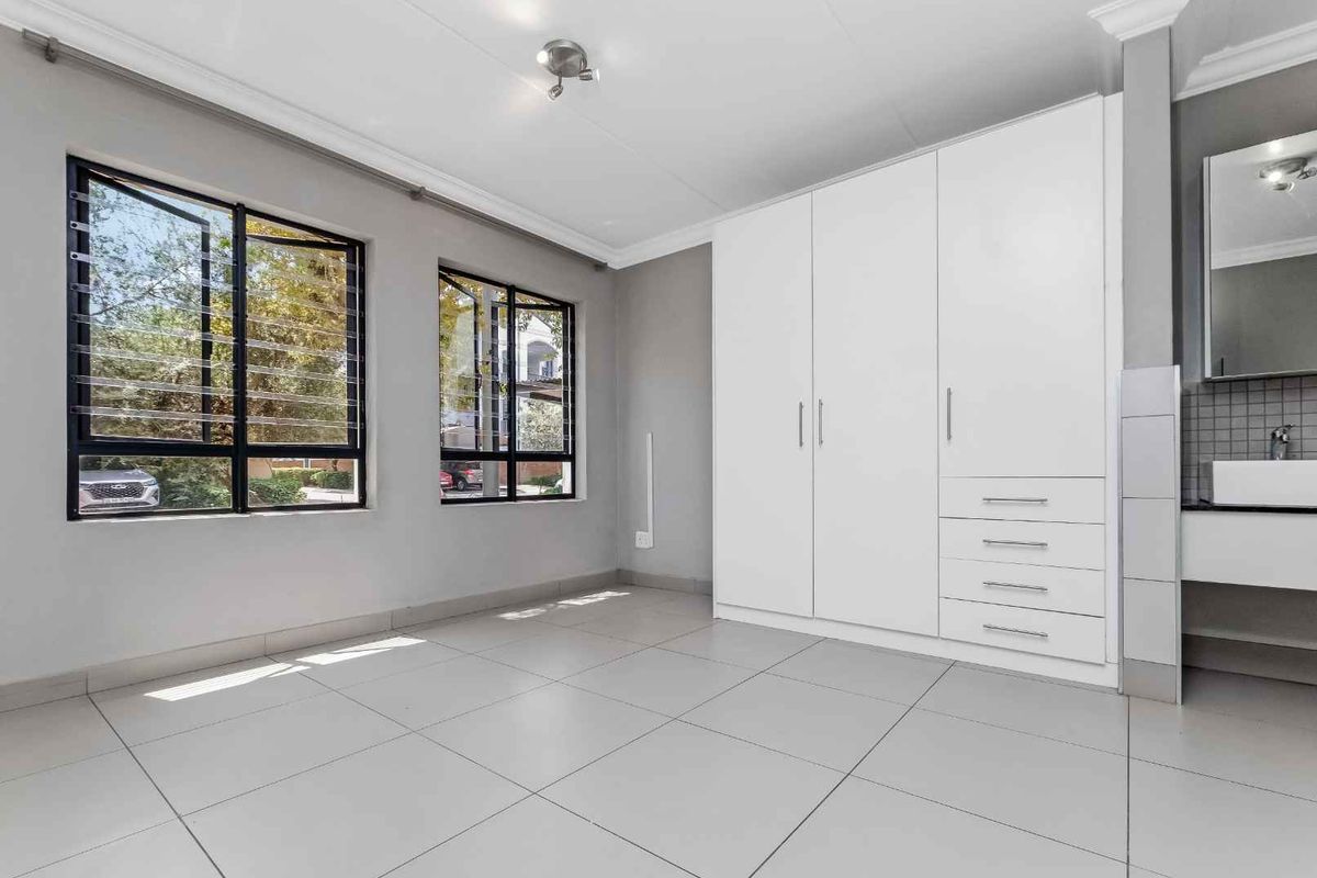 A bright bedroom featuring large windows, neutral tiled flooring, and a modern built-in wardrobe with integrated vanity and basin.