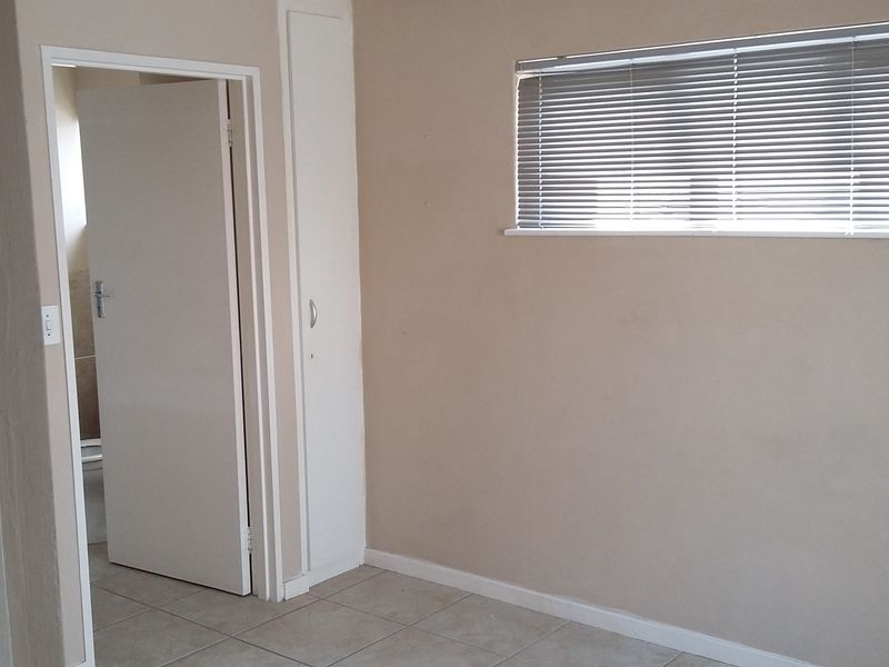 A clean, empty bedroom featuring neutral wall tones, tiled flooring, a built-in cupboard, and a window fitted with venetian blinds.