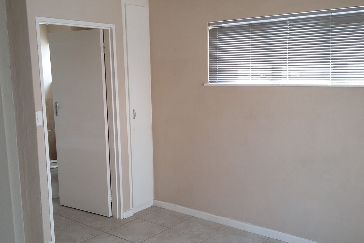 A clean, empty bedroom featuring neutral wall tones, tiled flooring, a built-in cupboard, and a window fitted with venetian blinds.