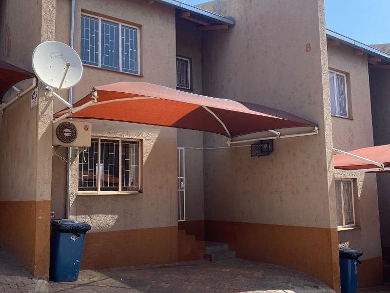 Front elevation of a townhouse featuring a paved driveway, shade sail, and exterior air conditioning unit.
