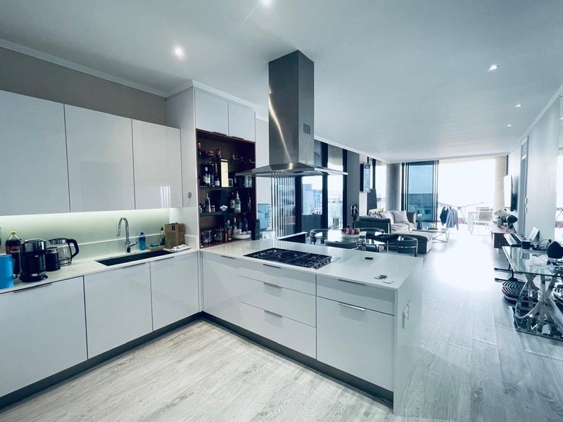 Modern open-plan kitchen featuring white cabinetry, a central island with gas cooktop, and integrated stainless steel extractor, flowing into a bright living area.