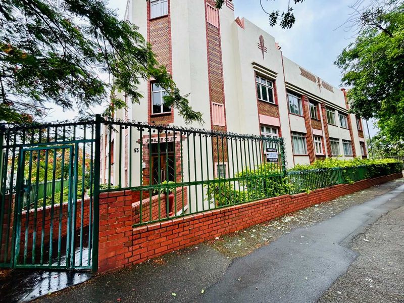 Street-level view of the Jubilee Court apartment building, featuring an art deco facade, brick boundary wall, and secure metal gated entrance.