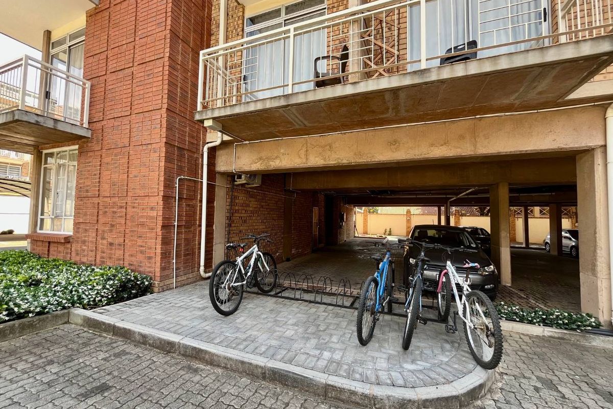 Ground level covered parking area beneath a residential balcony, featuring paved surfacing and brick facade construction.
