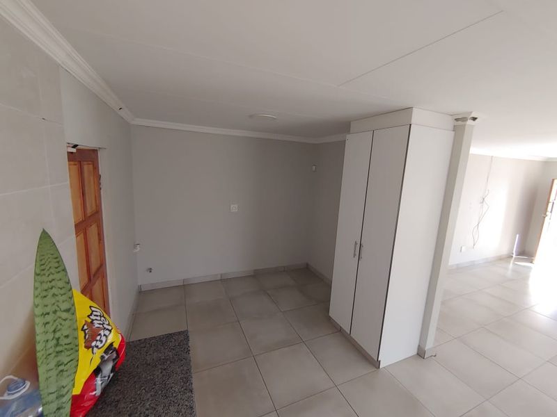 An open-plan living area featuring neutral tiled flooring, a built-in cupboard, and coved ceiling details, presenting a clean and versatile space.